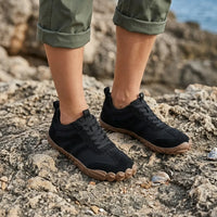 Zwarte barefoot schoenen gedragen door een model met opgerolde broekspijpen, staand op ruwe rotsen bij de zee. 20
