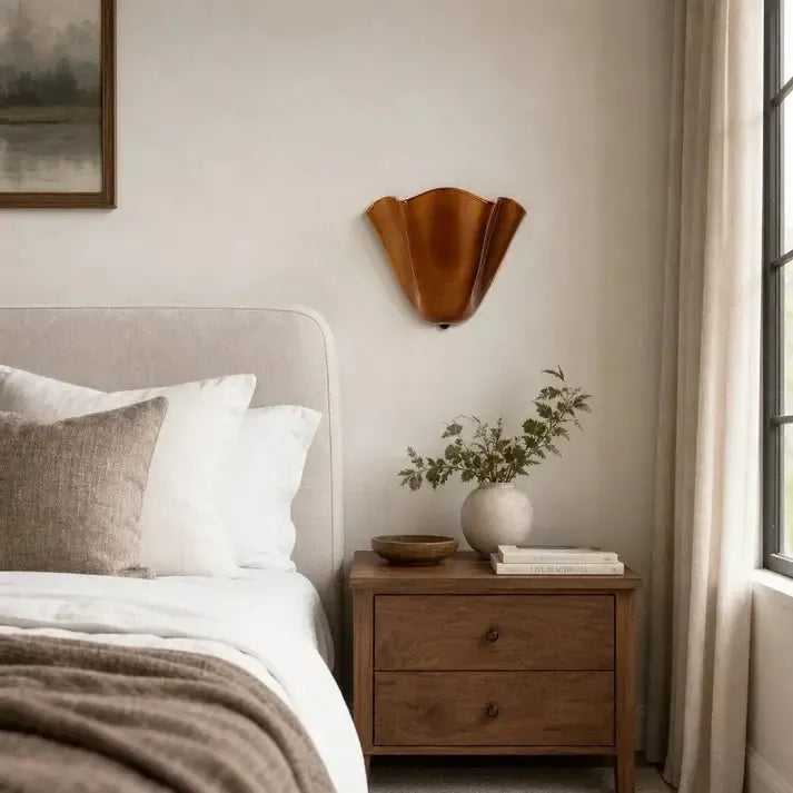 Uitgeschakelde bruine wandlamp met golvende rand boven een houten nachtkastje met vaas en boeken naast een bed. 3