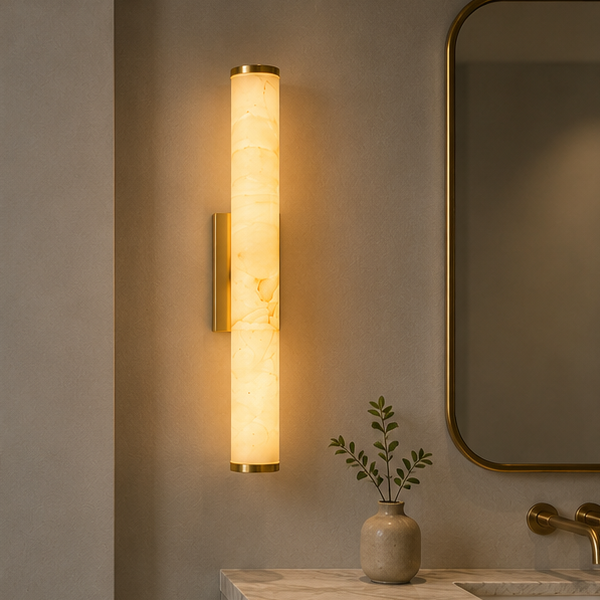 Verlichte verticale marmeren wandlamp met koperkleurige randen naast een gouden spiegel, boven een marmeren badkamerwastafel met vaasje. 2