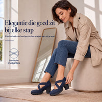 Vrouw in beige jas zit op een poef en trekt de marineblauwe schoen met gekruiste bandjes aan. 4
