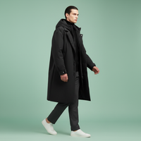 Model loopt in open zwarte lange jas met capuchon, grijze broek en witte sneakers tegen een muntgroene achtergrond. 7