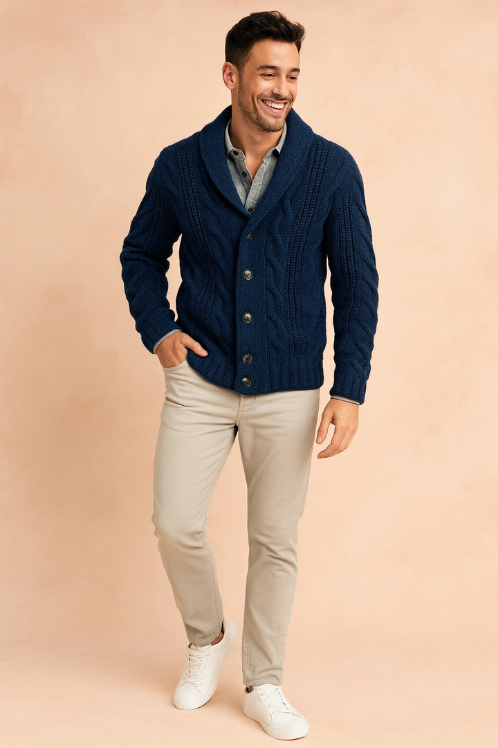 Volledig lichaamsbeeld van een man in het donkerblauwe kabelvest met beige broek en witte sneakers, voor een perzikkleurige achtergrond. 2