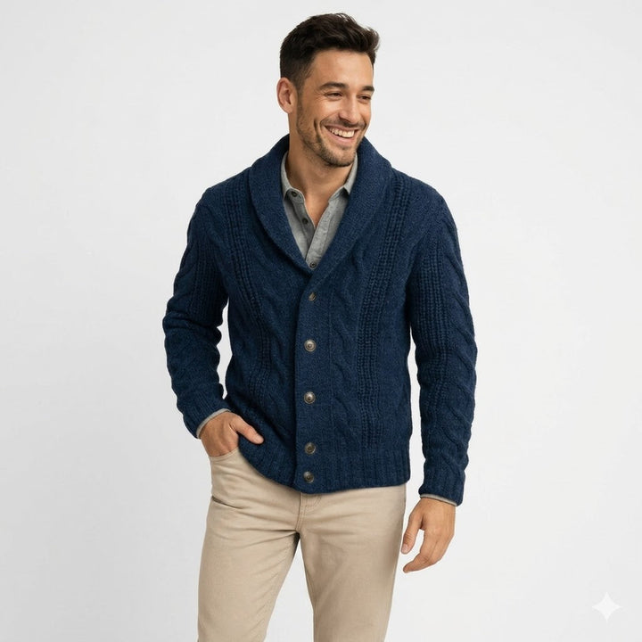 Lachende man op witte achtergrond draagt het donkerblauwe kabelvest met sjaalkraag, een hand in de zak van zijn beige broek, halfnaaldbeeld. 1