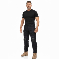 Man met baard staat in volledige lengte in zwart shirt, de zwarte cargobroek met kniekappen en beige laarzen. 7