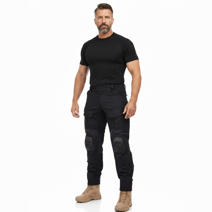 Man met baard staat in volledige lengte in zwart shirt, de zwarte cargobroek met kniekappen en beige laarzen. 7