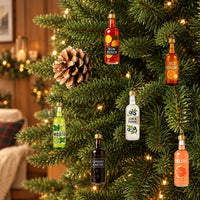 Zes gekleurde flesjes als kerstbal in een verlichte kerstboom, naast een dennenappel, met een gezellige woonkamer op de achtergrond. 1
