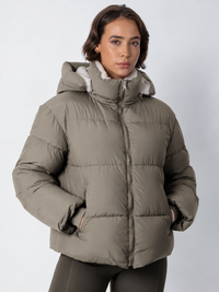 Model in taupekleurige pufferjack, dichtgeritst met capuchon op, met teddyvoering zichtbaar aan de kraag 18
