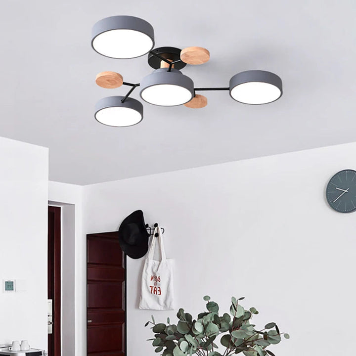 Grijze plafondlamp met vier ronde kappen en houten schijfjes aan zwarte armen, in een witte hal met kapstok, tas en klok. 5
