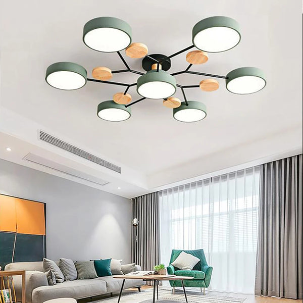 Groene plafondlamp met zeven ronde kappen en houten schijfjes aan zwarte armen, boven een woonkamer met grijze bank en groene fauteuil. 2