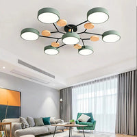 Groene plafondlamp met zeven ronde kappen en houten schijfjes aan zwarte armen, boven een woonkamer met grijze bank en groene fauteuil. 2