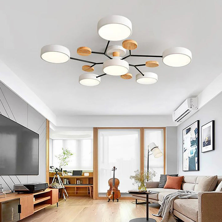 Witte plafondlamp met zes ronde kappen en houten schijfjes aan zwarte armen, hangend in een lichte woonkamer met televisie en beige bank. 3