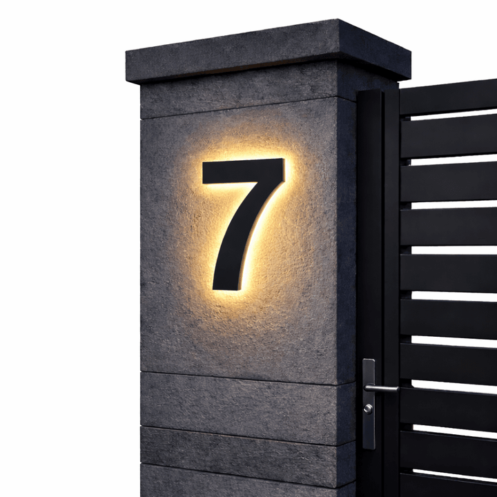 Zwart huisnummer 7 met warm achtergrondlicht op een grijze stenen pilaar naast een zwarte poort met horizontale latten. 13