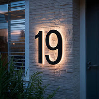 Verlicht zwart huisnummer 19 op een witte bakstenen gevel naast de voordeur, met planten op de voorgrond. 4