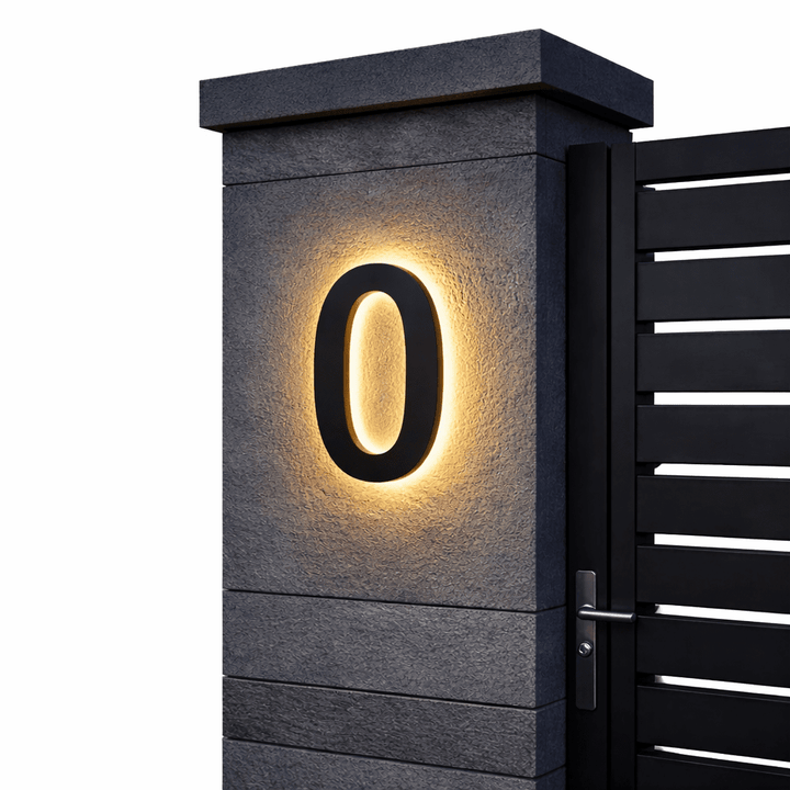 Zwart huisnummer 0 met warm achtergrondlicht op een grijze stenen pilaar naast een zwarte poort, op witte achtergrond. 6