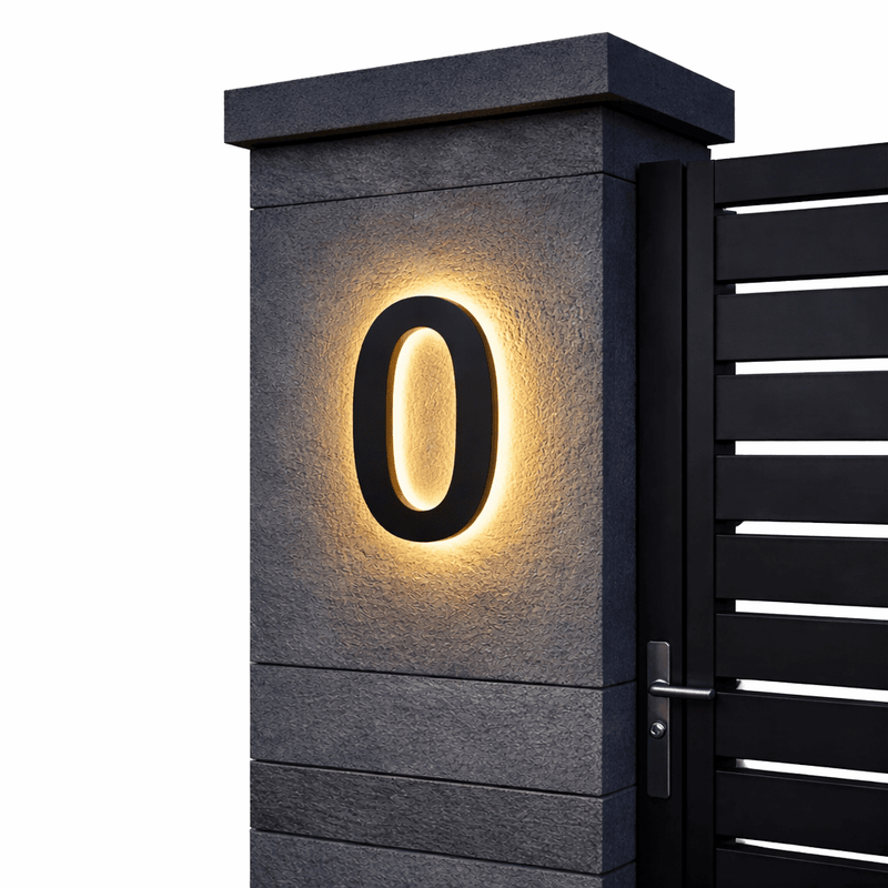 Zwart huisnummer 0 met warm achtergrondlicht op een grijze stenen pilaar naast een zwarte poort, op witte achtergrond. 6