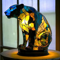 Zittende panter in blauw, groen en geel glas-in-loodmozaïek op een ronde voet, voor een raam. 16