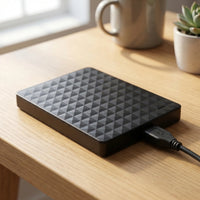 Zwarte harde schijf met ruitpatroon en aangesloten USB-kabel op een houten tafel naast een mok en plantje 1