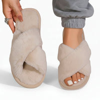 Beige pluche slippers met gekruiste banden; het model toont één slipper in de hand en draagt de andere bij een grijze joggingbroek. 7