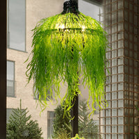 Verlichte kroonluchter behangen met lange, slierterige groene kunstplanten, hangend bij een raam met uitzicht op een stenen 2