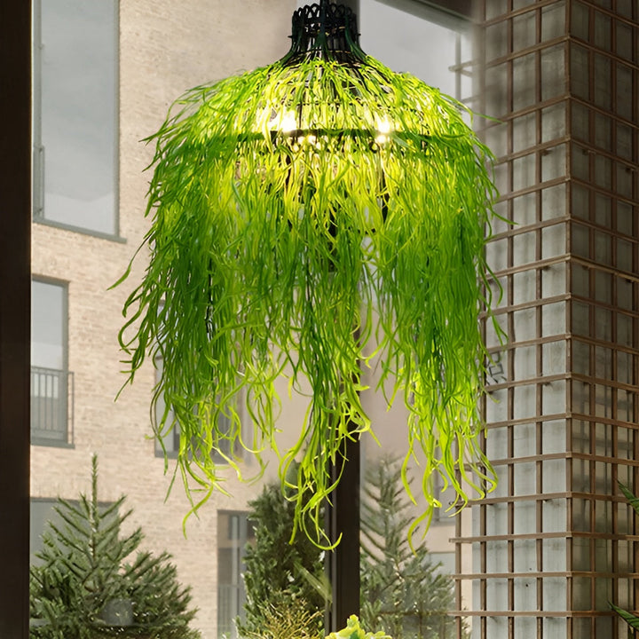 Verlichte kroonluchter behangen met lange, slierterige groene kunstplanten, hangend bij een raam met uitzicht op een stenen 2