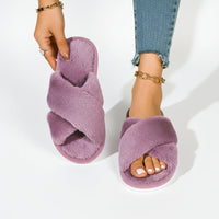 Paarse pluche slippers met gekruiste banden; het model houdt er één omhoog en draagt de andere bij een spijkerbroek. 8