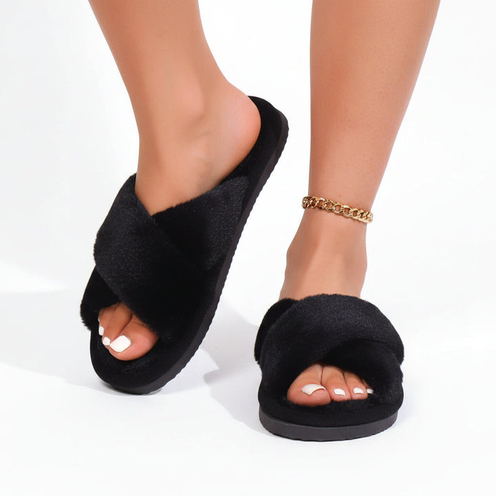 Zwarte pluche slippers met gekruiste banden aan de voeten van een model, met een gouden enkelbandje. 3