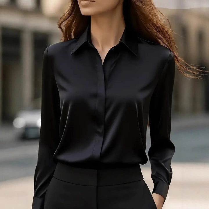 Model draagt de zwarte glanzende blouse met verdekte knoopsluiting, lange mouwen en zwarte broek, straat op de achtergrond 1