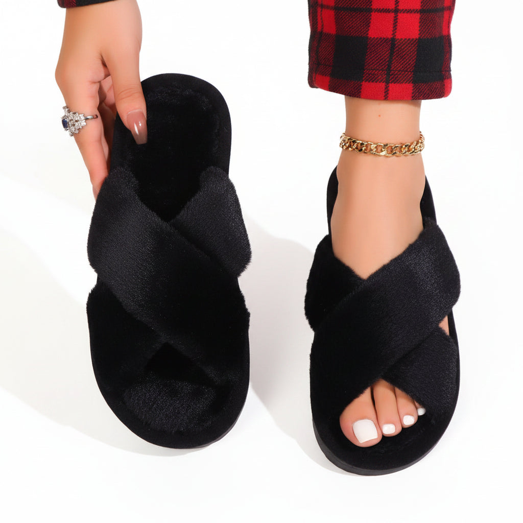 Zwarte pluche slippers met gekruiste banden; het model toont één slipper en draagt de andere bij een rood geruite broek. 9