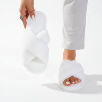 Witte pluche slippers met gekruiste banden; het model houdt er één omhoog en draagt de andere bij een grijze broek. 6