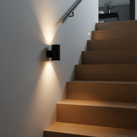 Zwart wandlampje aan een lichte muur naast een houten trap, met warme lichtbundels omhoog en omlaag. 5