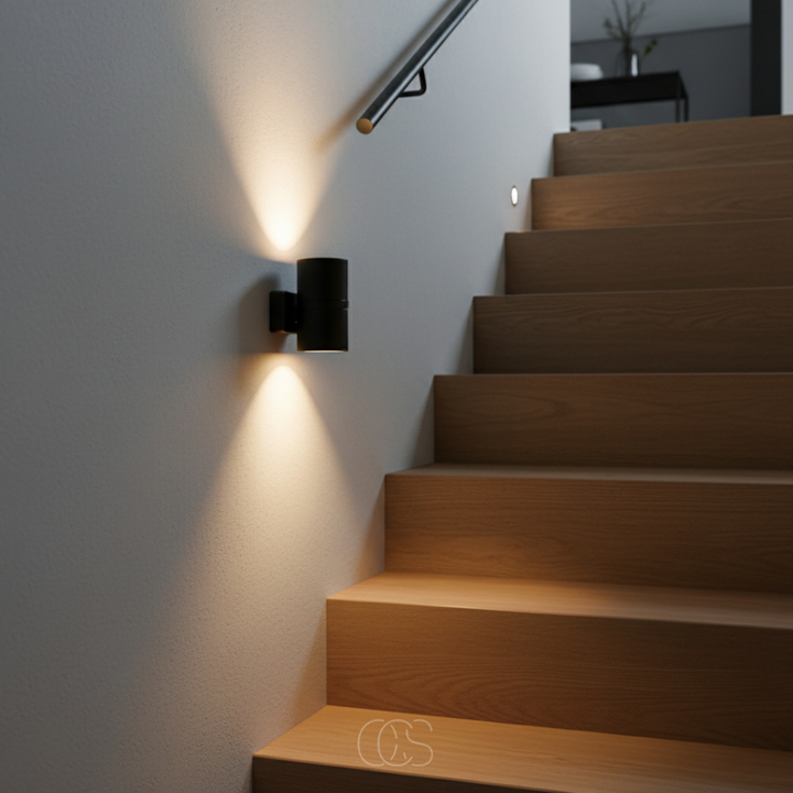 Zwart wandlampje aan een lichte muur naast een houten trap, met warme lichtbundels omhoog en omlaag. 5