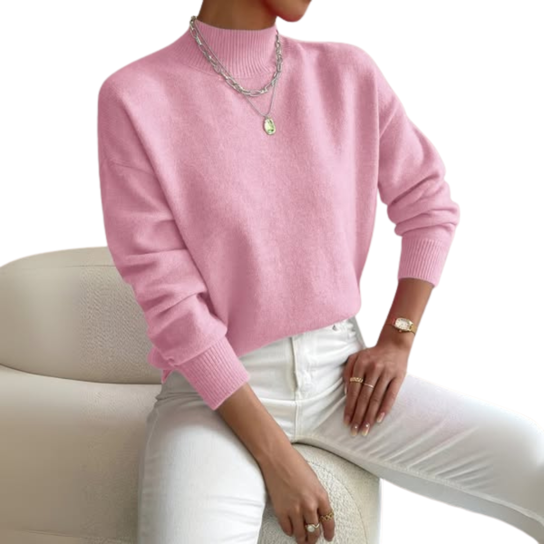 Model draagt de roze trui met ribboorden en lange mouwen, gecombineerd met witte broek en zilverkleurige kettingen, zittend op een crèmekleurige fauteuil. 1