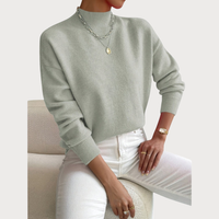 Model draagt de trui in saliegroen met ribkraag en boorden, met witte broek, gouden horloge en kettingen op een bouclé fauteuil. 2