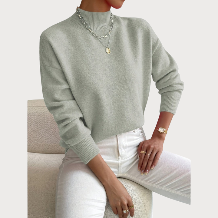 Model draagt de trui in saliegroen met ribkraag en boorden, met witte broek, gouden horloge en kettingen op een bouclé fauteuil. 2