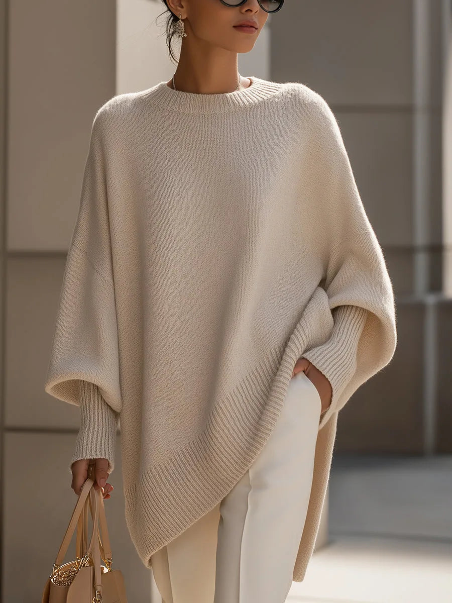 Model draagt de oversized trui in beige met witte broek, een hand in de zak en een tas in de andere. 1