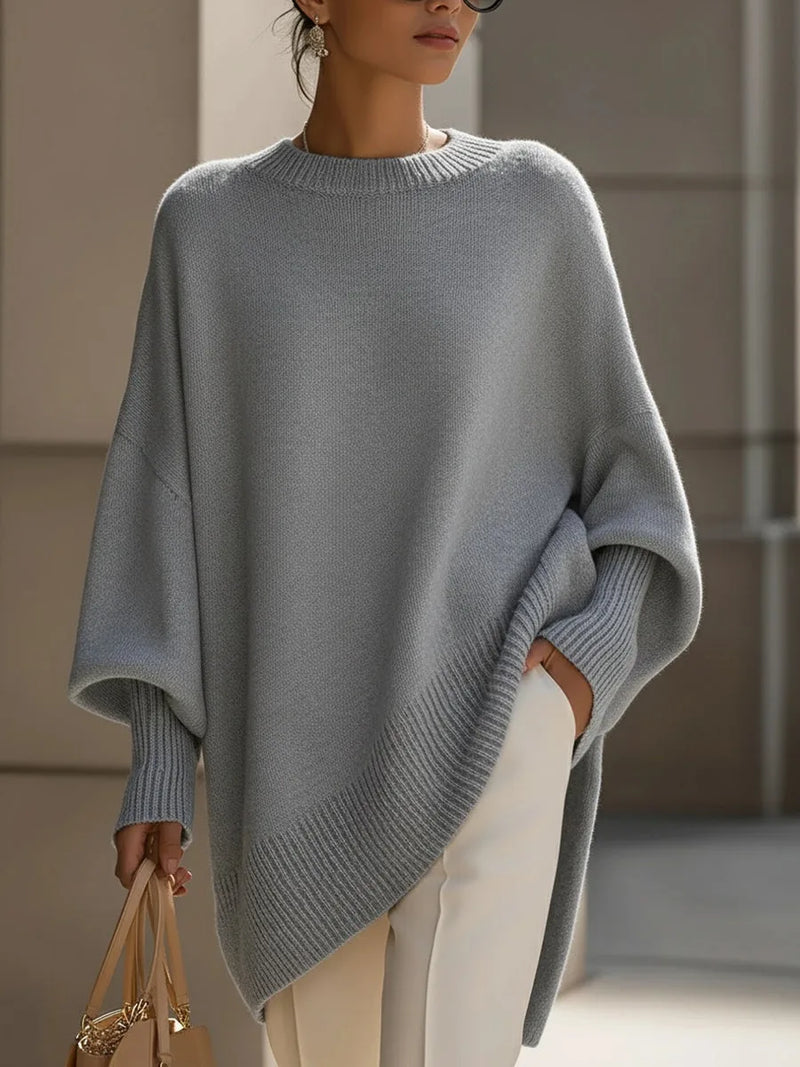 Model draagt de oversized trui in grijs met witte broek, een hand in de zak. 9
