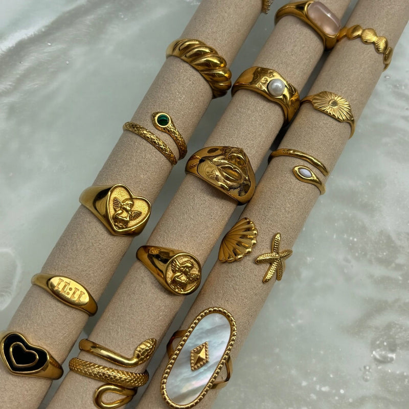 Verschillende gouden ringen op beige rollen, waaronder de hartring met engeltje, slangenringen, een parelring en een schelpring. 5