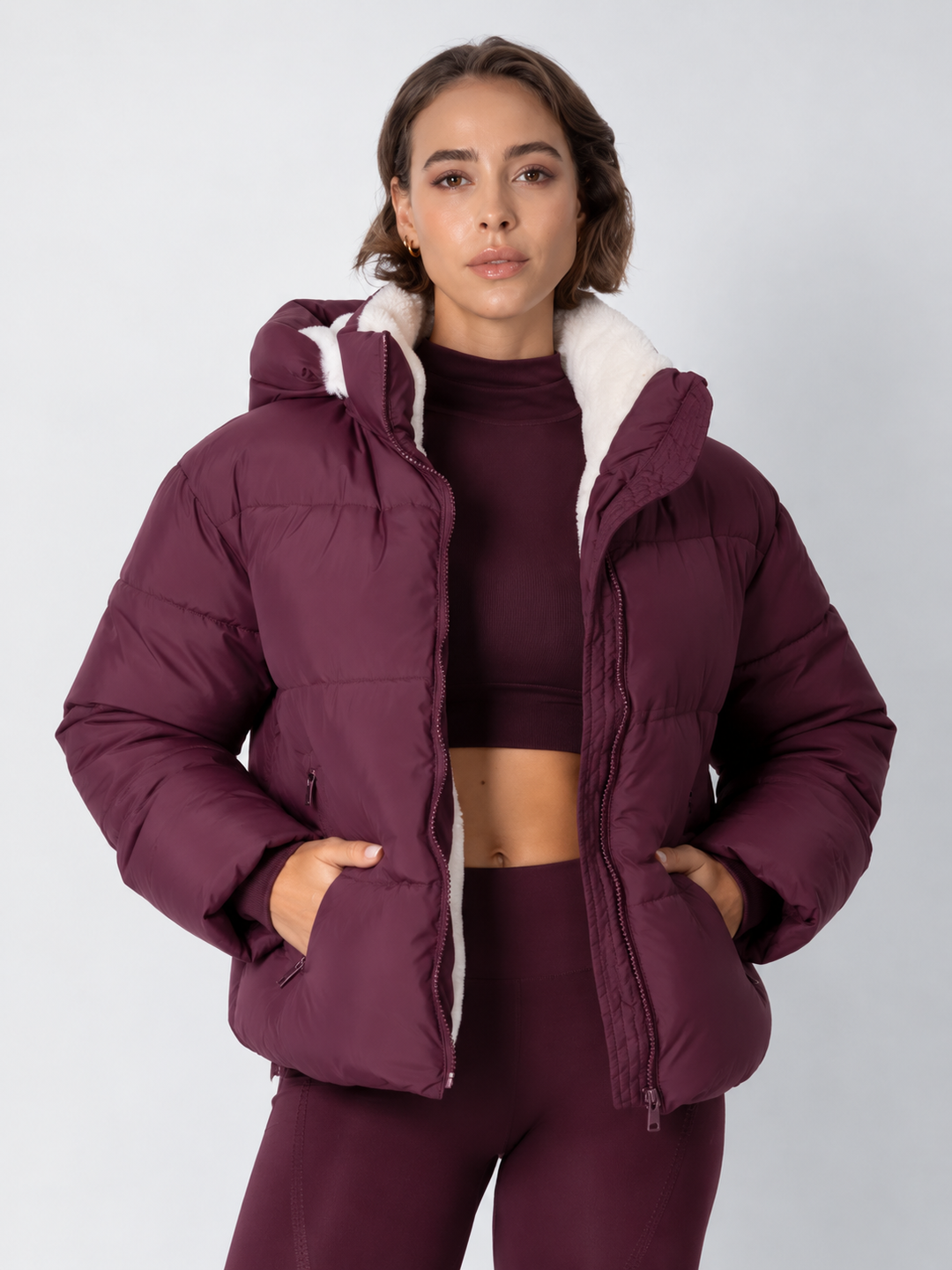 Model in bordeauxrode pufferjack open gedragen, met witte fleece voering en handen in de zakken 16