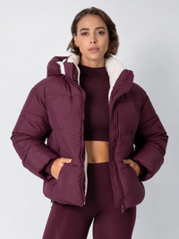 Model in bordeauxrode pufferjack open gedragen, met witte fleece voering en handen in de zakken 16