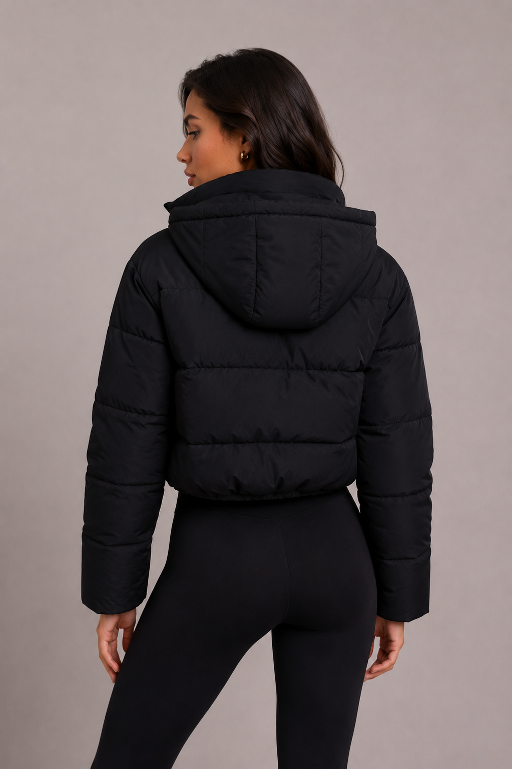 Achterkant van de zwarte gewatteerde jas met horizontale stiksels, hoge kraag en capuchon, gedragen door een model in zwarte legging. 4