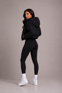 Model kijkt over haar schouder en toont de zijkant en achterkant van de zwarte pufferjas met capuchon, met legging en witte sneakers. 5