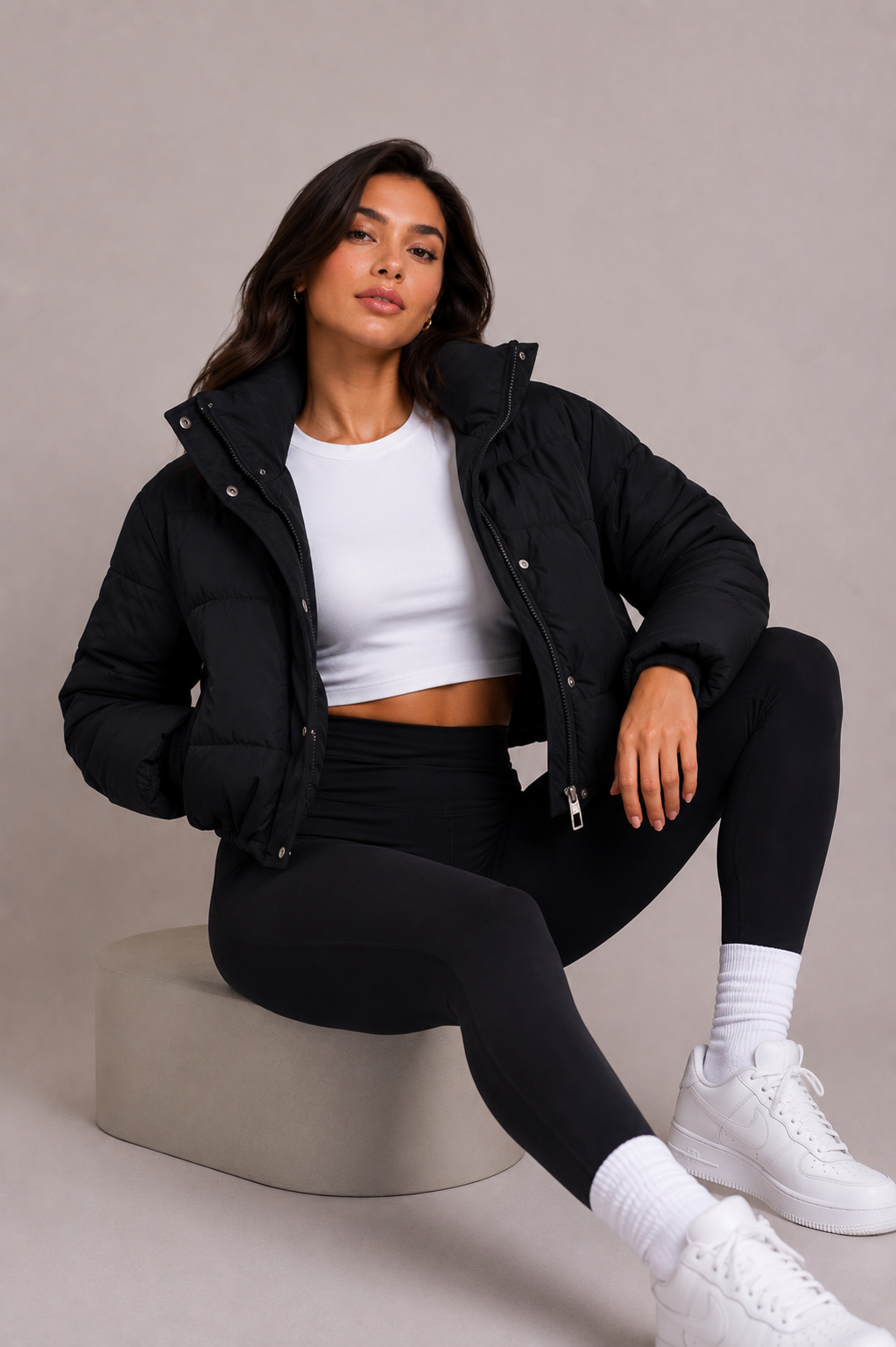 Zittend model draagt de zwarte gewatteerde jas open met wit topje, zwarte legging en witte sneakers, poserend op een neutraal blok. 6