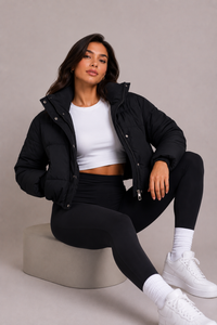 Zittend model draagt de zwarte gewatteerde jas open met wit topje, zwarte legging en witte sneakers, poserend op een neutraal blok. 6