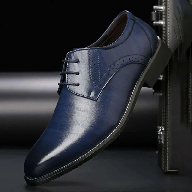 Blauwe glanzende veterschoen met brogue-perforatie, schuin tegen een donkere koffer geplaatst. 5
