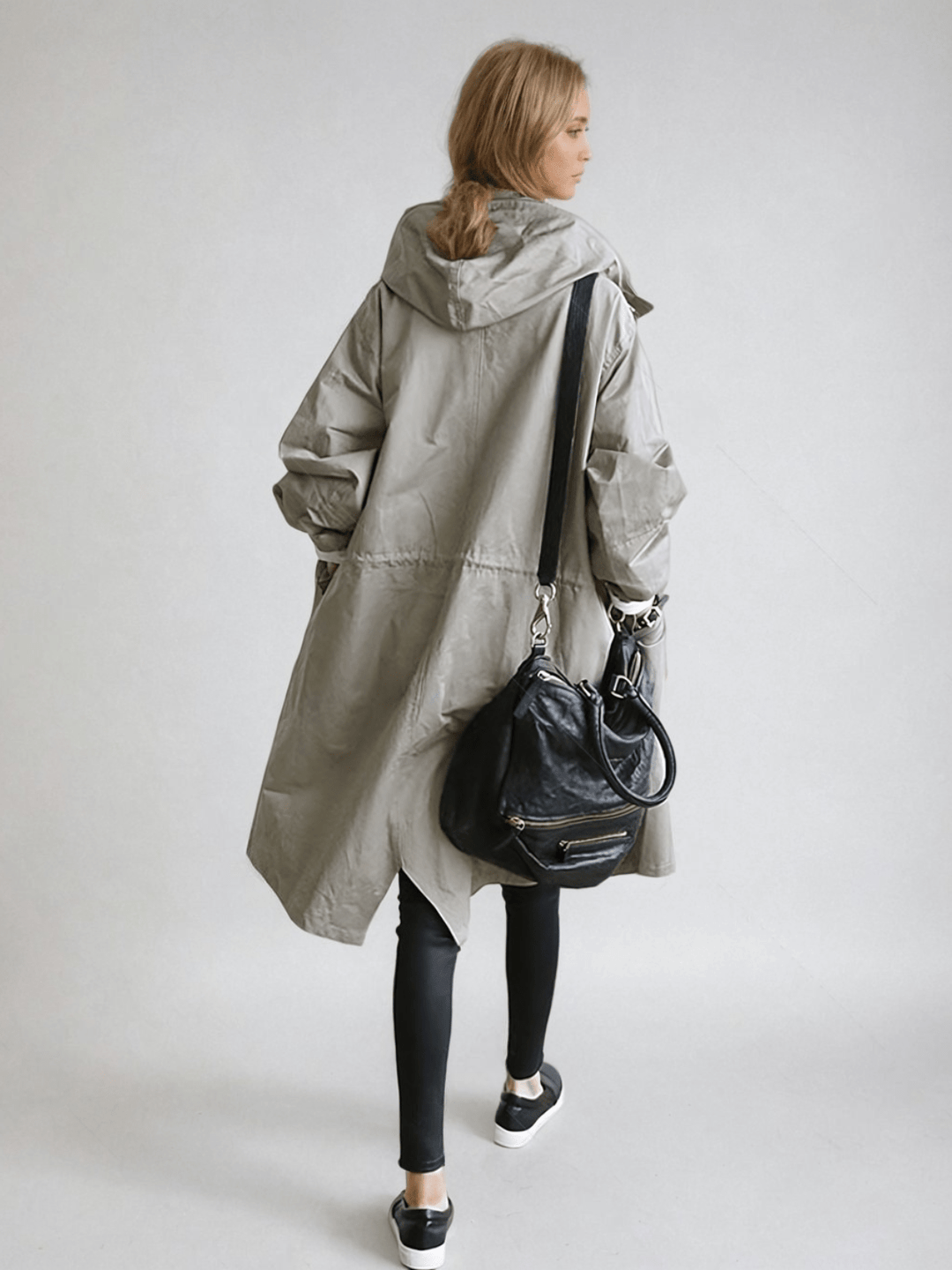 Achterkant van de grijze trenchcoat op een model, met capuchon, taillekoord en een zwarte schoudertas aan de arm. 4