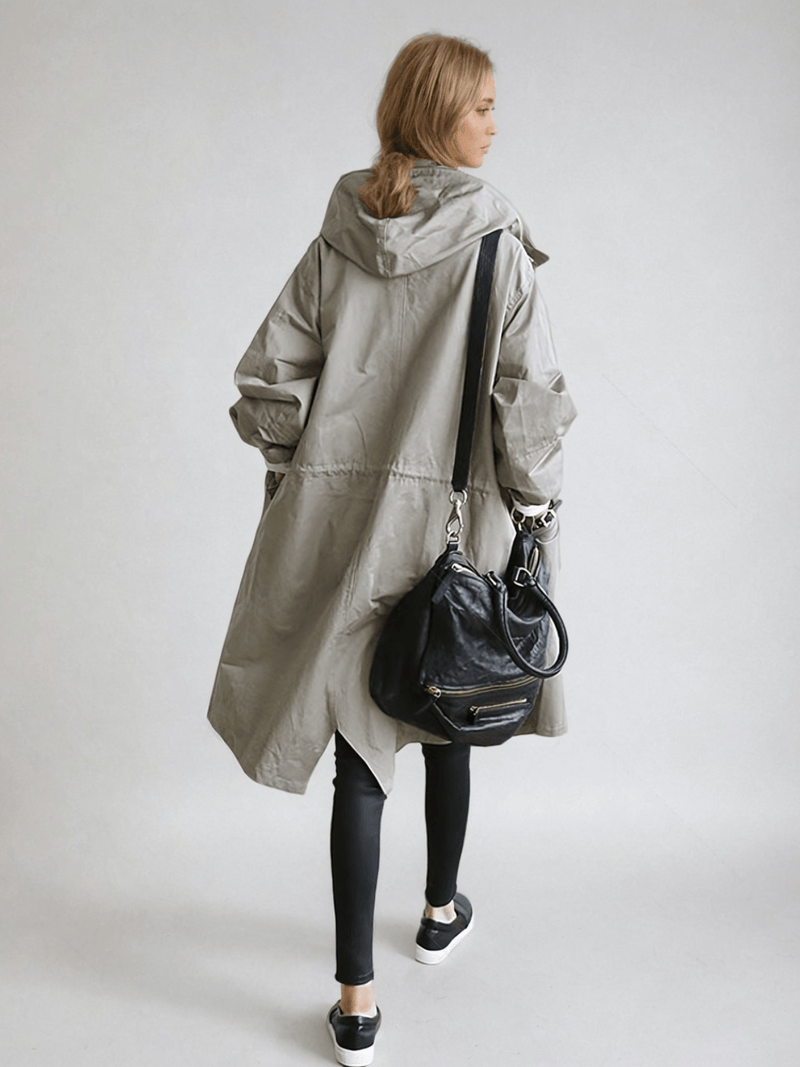 Achterkant van de grijze trenchcoat op een model, met capuchon, taillekoord en een zwarte schoudertas aan de arm. 4