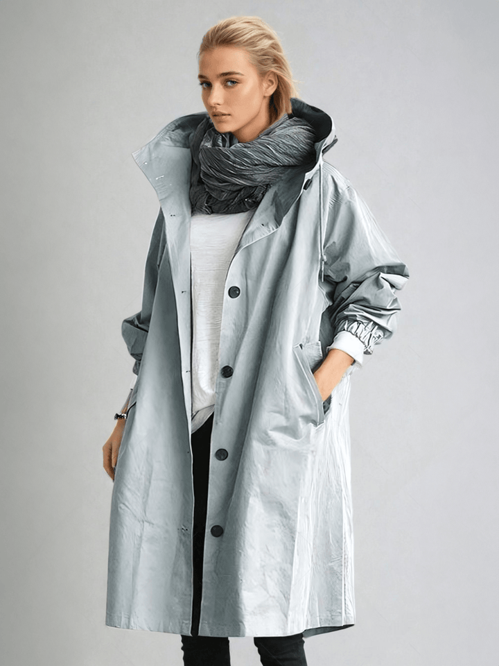 Model draagt de lichtgrijsblauwe trenchcoat met glanzende stof, capuchon en knopen, met een hand in de zak. 5