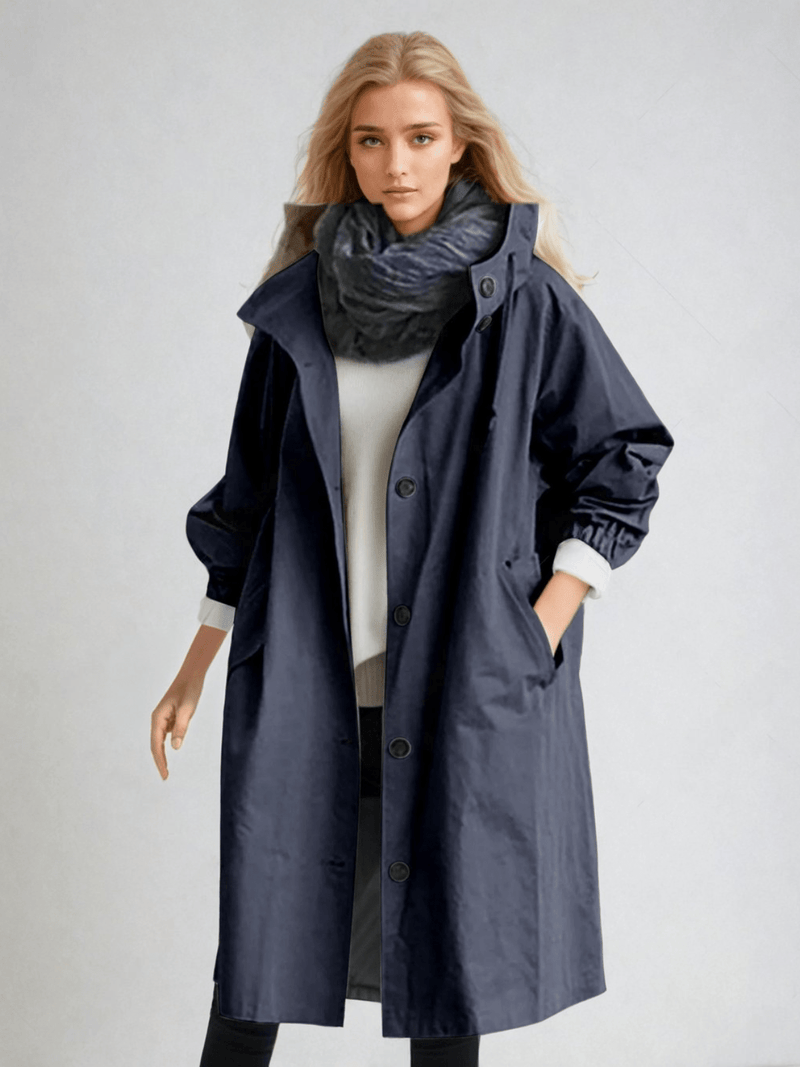 Model draagt de donkerblauwe trenchcoat met grote capuchon en knopen, gecombineerd met een witte trui en donkere sjaal. 7