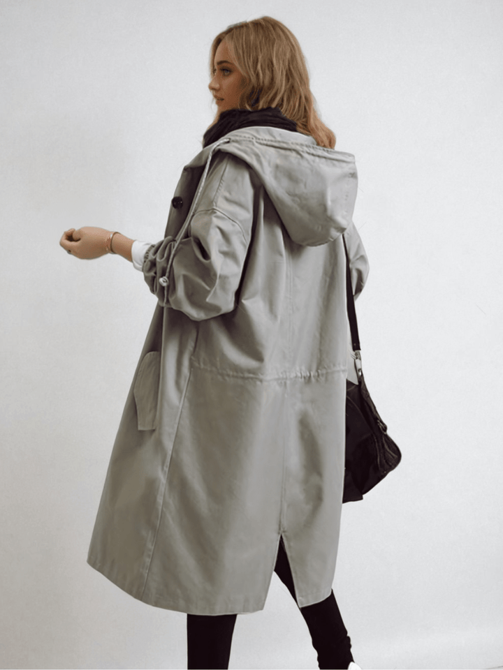 Achterzijde van de grijze trenchcoat in beweging, met capuchon, trekkoord in de taille, zijsplit en een zwarte tas. 8