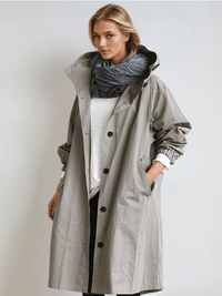 Model draagt de grijze trenchcoat met capuchon open over een wit shirt en donkere sjaal, met een hand in de zak. 1